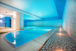 Piscina coperta per famiglie – Family Hotel Polsa, Brentonico Piscina coperta riscaldata del family hotel Polsa in Trentino, con vasca dal profilo curvo, pareti in pietra chiara e luci soffuse